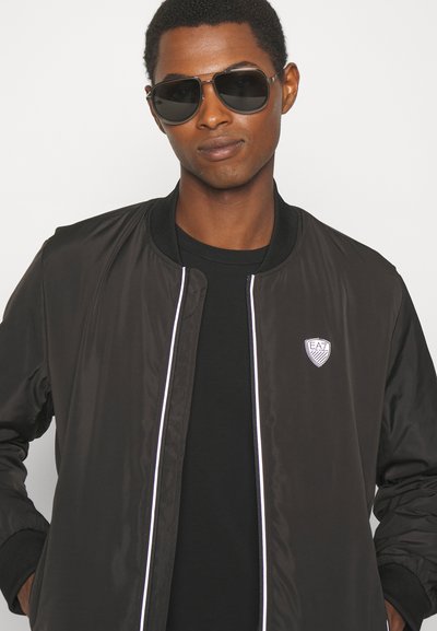 Veste bomber noire avec des passepoils blancs, arborant un logo sur la poitrine. Portée sur une chemise noire ; associée à des lunettes de soleil foncées. Texture lisse.
