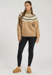 Brun stickad tröja med hög krage, prydd med vita och svarta geometriska mönster längs jägen; tillsammans med svarta leggings och bruna tofflor.