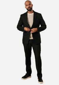 Blazer nero sopra una camicia a righe, abbinato a pantaloni neri su misura e sneakers nere. Dettagli notevoli includono i bottoni sul blazer e una vestibilità classica.