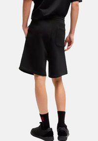Czarne sportowe szorty o luźnym kroju, wykonane z miękkiej tkaniny, z tylną kieszenią i elastycznym pasem. Noszone z czarnymi butami i skarpetkami.