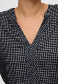 Blouse en pied-de-poule navy et gris présentant un décolleté en V, une texture douce et une coupe décontractée. Le tissu présente une variation de motif subtile.