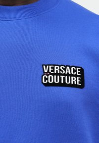 Detailný záber modrého mikiny s čiernym a bielym logo nášivkou "Versace Couture" na hrudi, s malým ružovým textom "Jeans".