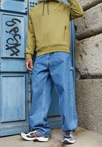 Sudadera con capucha verde oliva con un bolsillo frontal con cremallera, combinada con jeans azules holgados de mezclilla. El conjunto se completa con zapatos deportivos negros y plateados.