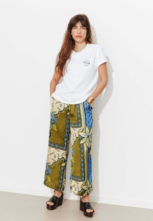 Femme aux longs cheveux bruns portant un t-shirt blanc, un pantalon large à motifs verts et bleus, des sandales à plateforme, des bracelets en or et un collier.