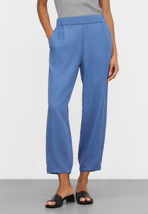Femme portant un pantalon bleu ample avec poches latérales et des sandales noires à talons carrés ouverts, debout devant un fond uni.