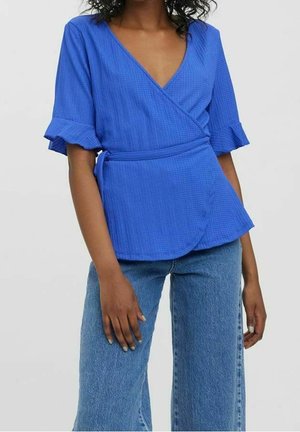 Femme portant un chemisier cache-cœur bleu vif avec des manches courtes à volants et un jean taille haute bleu clair sur un fond uni.