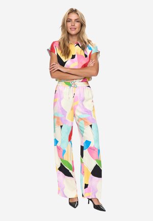 Kobieta stojąca z założonymi ramionami, ubrana w kolorową koszulę z abstrakcyjnym wzorem na krótkim rękawie oraz dopasowane szerokie spodnie i czarne obcasy.
