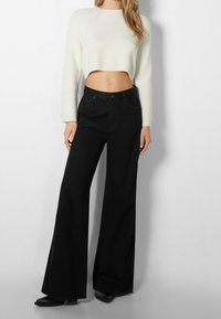 Schwarze weite Hosen mit Fünf-Taschen-Design, Knopfverschluss und genähten Details, kombiniert mit einem kurzärmeligen, cremfarbenen Pullover. Sanfte Textur.