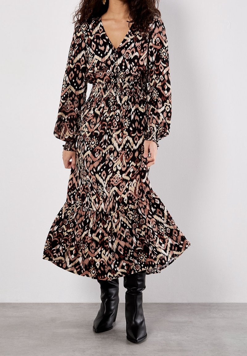 Femme portant une robe midi à manches longues avec un motif, un col en V et une taille froncée, associée à des bottes noires à talons hauts.