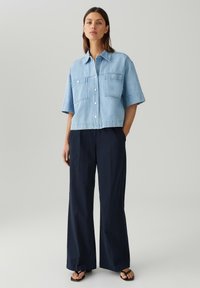 Lichtblauwe denim cropped blouse met korte mouwen, voorzakken en knoopsluiting, gecombineerd met donker marineblauwe wijde broek en sandalen.