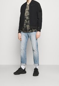 Svart bomberjacka med dragkedja, grått tie-dye t-shirt, ljusblå jeans och svarta sneakers med vita detaljer. Avslappnat outfit.