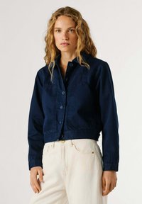 Blondynka o kręconych włosach, ubrana w granatową kurtkę na guziki i białe spodnie z wysokim stanem, stoi na tle jednolitego tła.