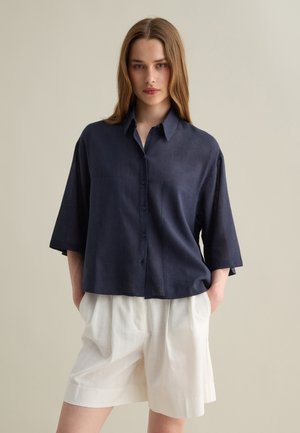 Navy blue short-sleeve linen shirt with a button-up front, two chest pockets, and a relaxed fit, paired with white pleated shorts.