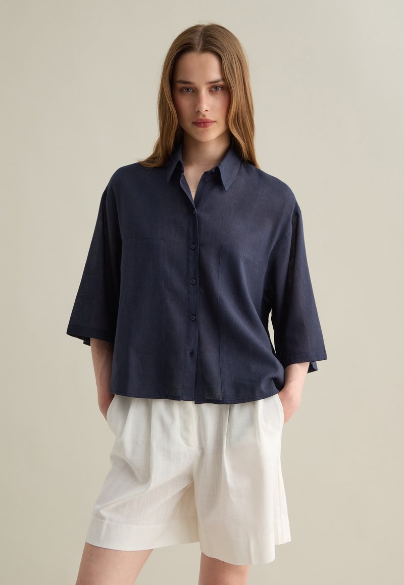 Navy blue short-sleeve linen shirt with a button-up front, two chest pockets, and a relaxed fit, paired with white pleated shorts.