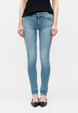 Inimene, kes kannab helesiniseid kitsaid teksaseid ja musti terava ninaga kontsakingi, seisab tavalise valge tausta ees.