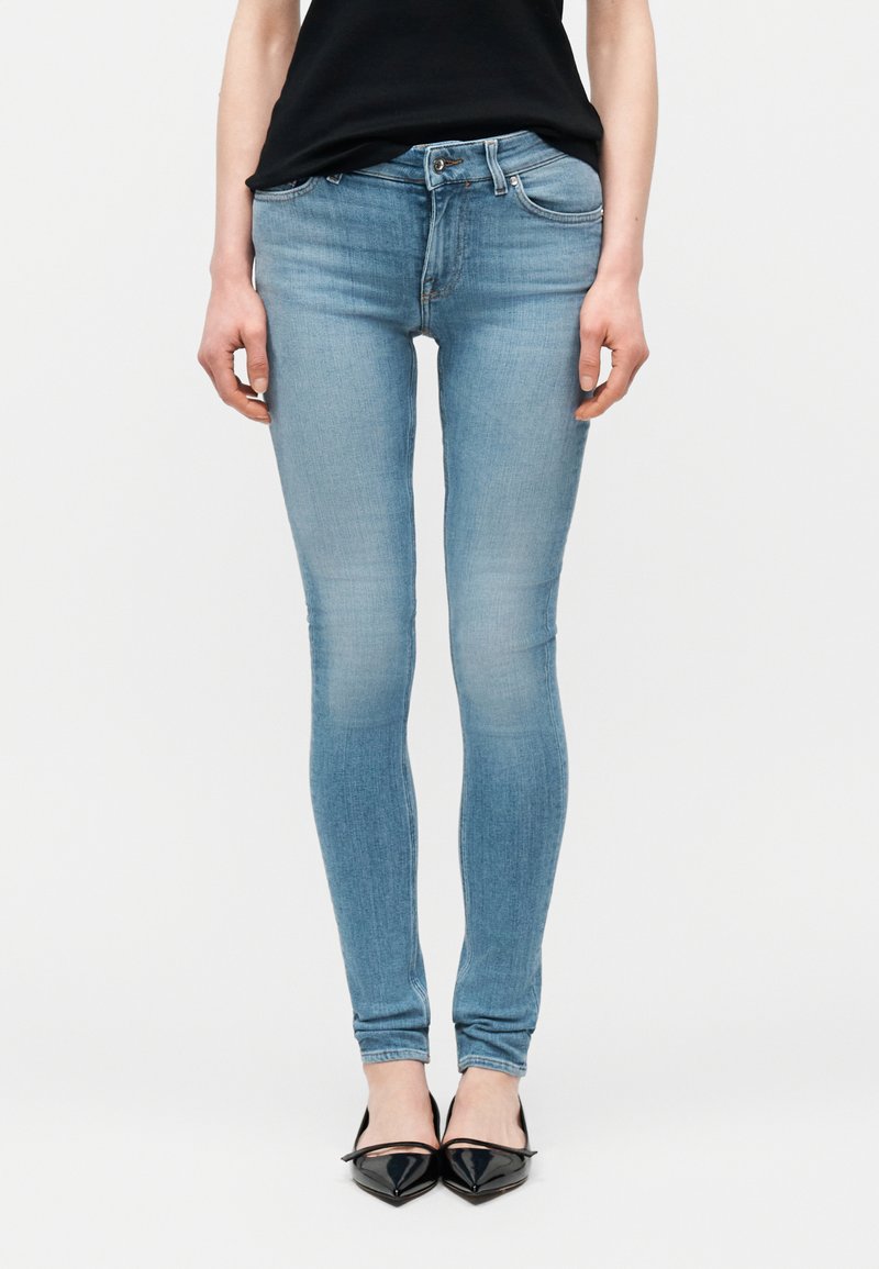 Άτομο που φορά στενά τζιν ανοιχτού μπλε χρώματος συνδυασμένα με μαύρα παπούτσια με μυτερή μύτη, στέκεται μπροστά σε απλό λευκό φόντο.