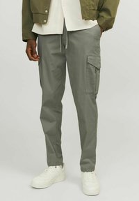Pantalones cargo gris claro con bolsillos laterales, confeccionados en una tela suave. Presentan un diseño ajustado y una cintura con cordón, combinados con zapatillas blancas.