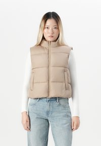 Ung kvinne som har på seg beige puffvest over hvit langermet topp og lyseblå jeans, står foran en enkel hvit bakgrunn.