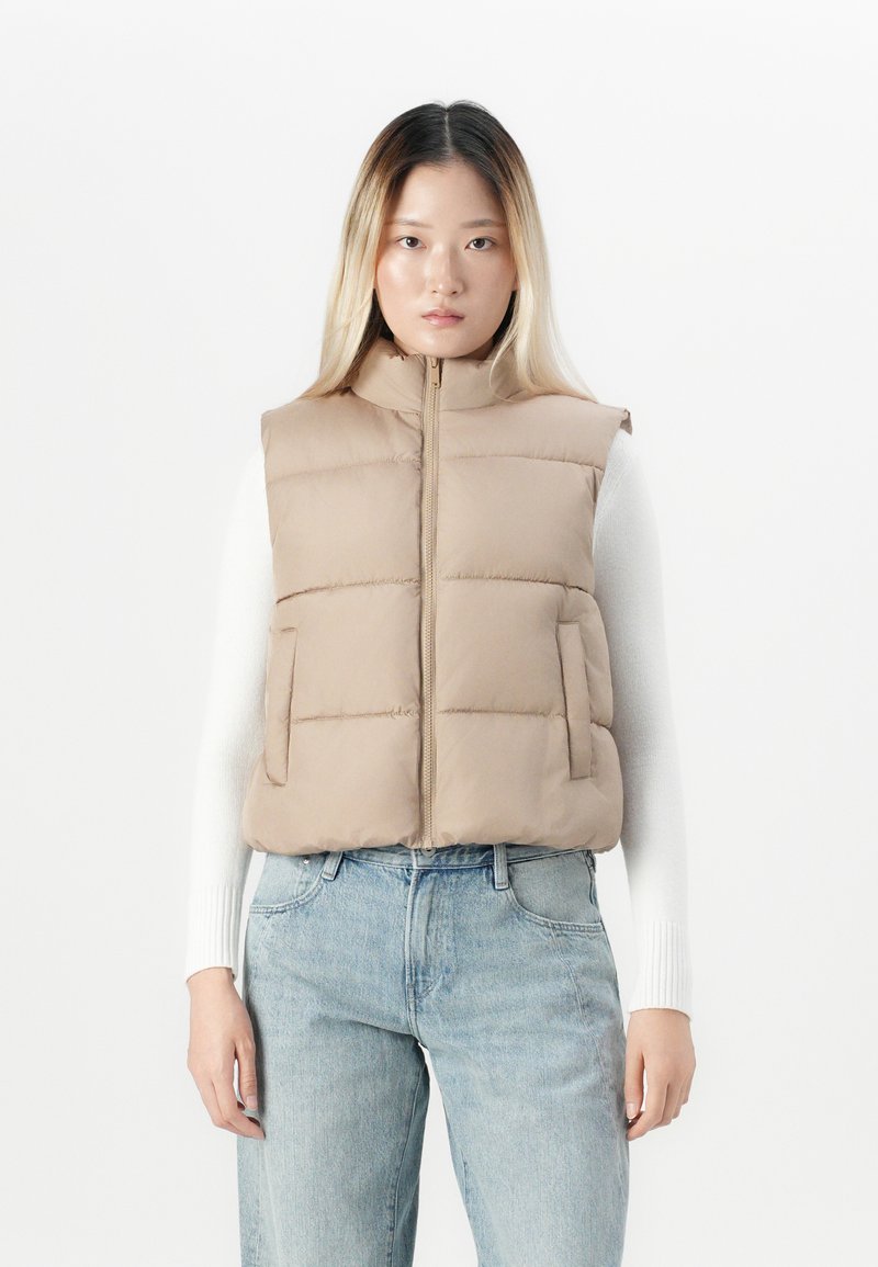 Ung kvinne som har på seg beige puffvest over hvit langermet topp og lyseblå jeans, står foran en enkel hvit bakgrunn.