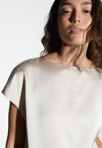 Top in satin color crema con scollo a barca e maniche corte. Presenta una texture liscia e cuciture minime per un aspetto elegante.