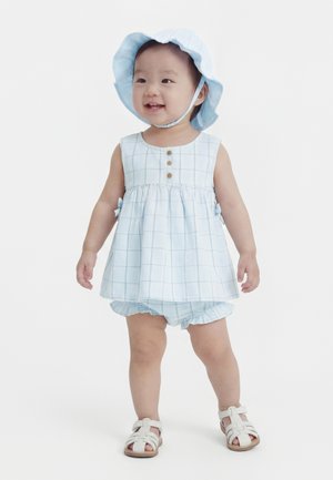 Copil mic purtând o rochie cu carouri albastre deschise, pălărie de soare asortată și sandale albe, stând în picioare și zâmbind pe un fundal simplu.