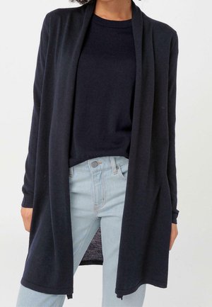 Personne portant un long cardigan noir ouvert sur le devant, par-dessus un haut noir et un jean bleu clair, sur un fond uni.
