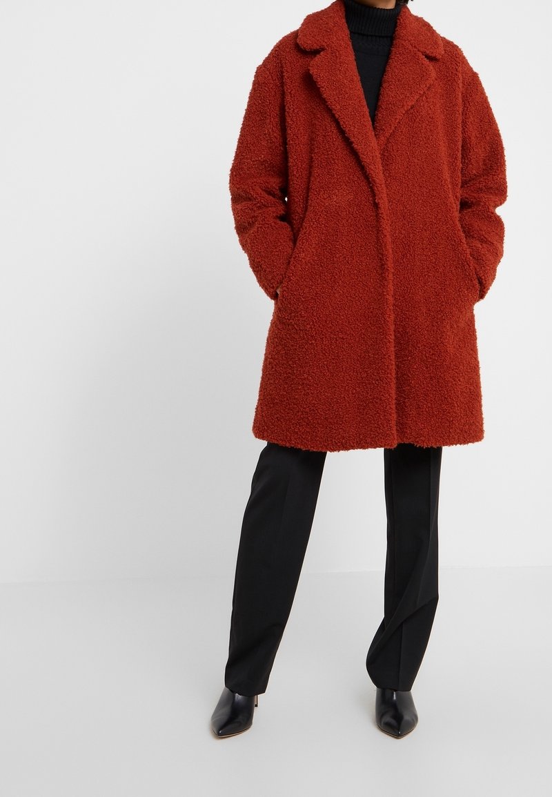 Rode textuur teddy coat met een gesneden kraag, voorzien van zijzakken en een relaxte pasvorm, gecombineerd met zwarte broeken en puntige laarzen.