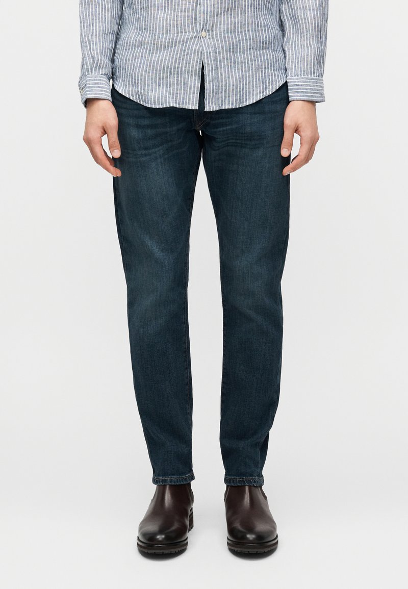 Άνδρας που φοράει σκούρο μπλε τζιν, καφέ δερμάτινα μπότες και ριγέ πουκάμισο μπλε και άσπρο, στέκεται με τα χέρια χαλαρά δίπλα στο σώμα.