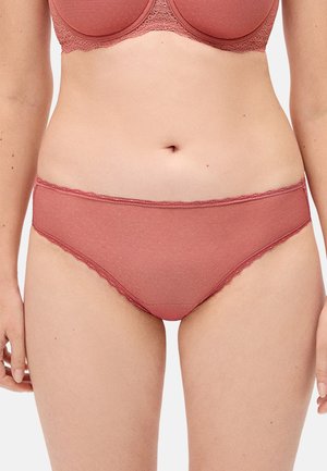 Mujer vistiendo un conjunto a juego de sujetador y ropa interior en color rosa empolvado, centrada en el torso y las caderas contra un fondo neutro.