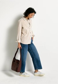 Beige corduroy overhemd met knopen, blauwe wijde jeans en witte sneakers. Bruine handtas met schouderband. Eenvoudige, casual outfit.