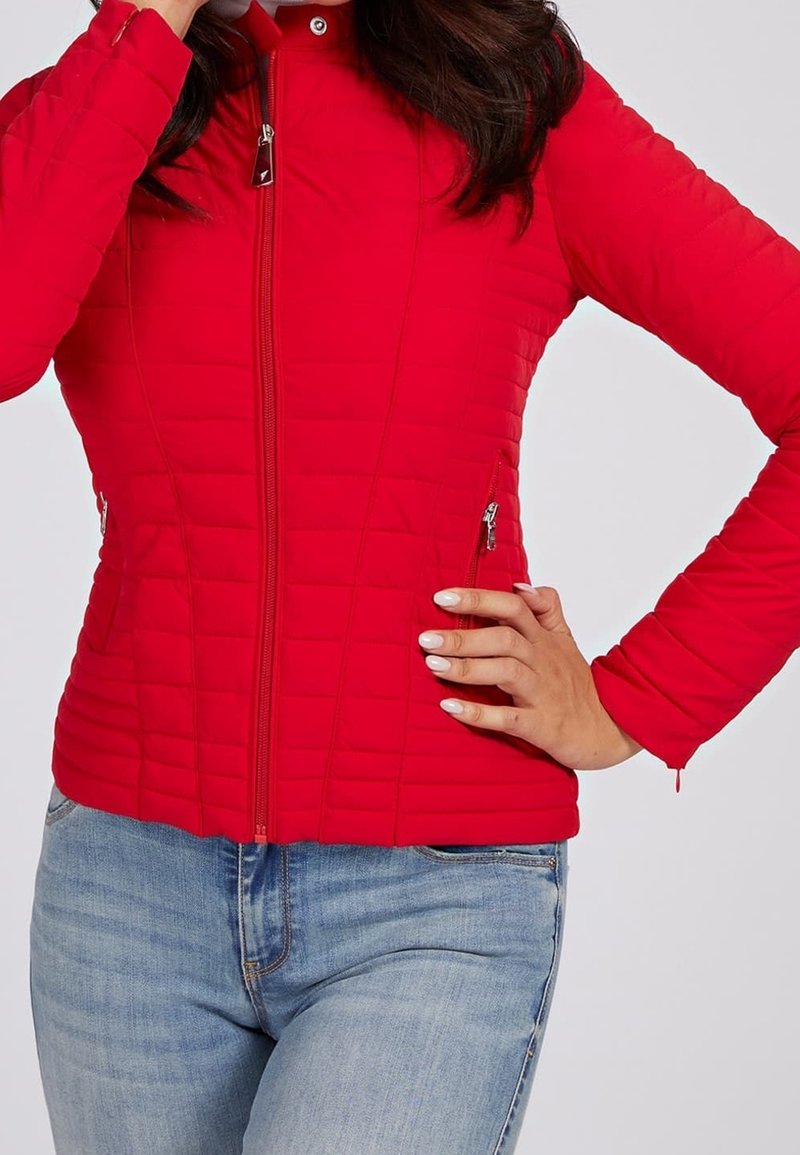 Femme portant une veste matelassée rouge ajustée avec des poches à fermeture éclair, posant avec une main sur la hanche et l'autre près du visage, associée à un jean bleu clair.