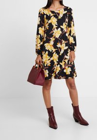 Vestido negro con estampado floral amarillo, de mangas largas y corte holgado. Combinado con un bolso burdeos y botas de tobillo de cuero texturizado.