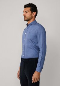 Chemise bleu clair à manches longues et col évasé, avec un petit logo brodé. Associée à un pantalon sombre. Texture lisse.