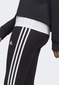 Svarta träningsleggings har tre vita vertikala ränder på sidan och en liten vit Adidas-logotyp. Tyget verkar slätt och elastiskt.