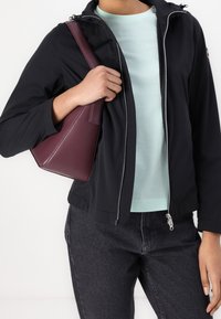 Svart jacka med dragkedja, ljus mintgrön skjorta under, matchad med mörka jeans och en vinröd triangulär väska i handen.