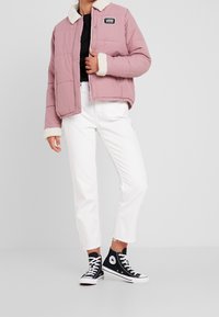 Lyserød polstret jakke med hvid pelskrave og manchetter, udstyret med trykknapper foran og Vans-logo. Bæres med hvide jeans og sorte høje sneakers.