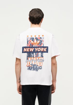 Person iført en hvid T-shirt med farverige "New York" og "NYC" grafik, inklusive stjerner og et trekløver-logo, set bagfra.