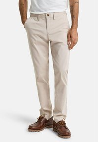 Chinos de algodón beige con corte recto, con cierre de botón, bolsillos laterales y una textura suave, combinados con zapatos marrones.