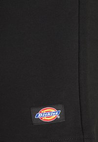 Černá tričkovina s žebrovanou texturou. Na spodním levém rohu je přišitá barevná štítek s logem "Dickies".