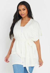 Vit, lätt cardigan med korta ärmar, ett knyt i midjan och en mjuk textur. Har en djup V-halsringning och öppen frontdesign.