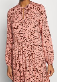 Růžové šaty s dlouhými rukávy, stahovacím límcem a černými skvrnitými vzory. Volný střih s nařaseným pasem, vyrobené z lehké látky.