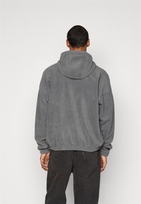 Šedá fleecová mikina s kapucí na šňůrku, spadlými rameny a ribovaný lem. Měkká textura, volný střih, kombinováno s tmavými džíny.