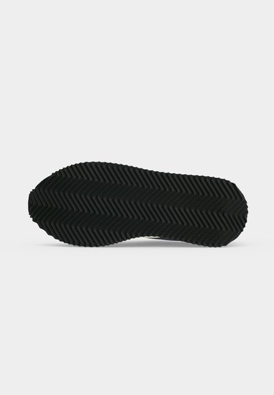 Semelle de chaussure noire avec un profond motif en zigzag façon chevrons, présentée sur fond blanc uni, mettant en avant la conception pour l’adhérence.