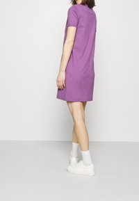 Fioletowa sukienka na krótki rękaw wykonana z bawełny, do kolan, o luźnym kroju. Noszona z białymi sneakersami i skarpetkami do kostek. Brak widocznych wzorów.