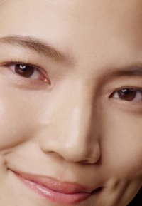 Gros plan d'un visage avec une peau lisse et claire, des yeux marron, des sourcils droits et foncés, et un sourire subtil révélant des lèvres pleines.