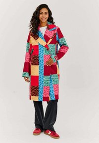 Farebný patchworkový kabát s odvážnymi vzormi v červenej, ružovej, zelenej a modrej farbe, so vzormi leopardov a geometrickými tlačami. Dvojradové zapínanie.