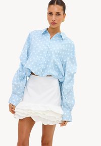 Femme portant une chemise bleue à pois ample et une jupe blanche courte à volants, debout contre un fond blanc uni.