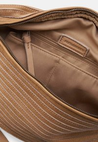 Interno di una borsa a tracolla in pelle marrone con una consistenza liscia, dotata di una tasca con zip e un'etichetta di marca, arricchita da cuciture diagonal.
