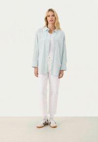 Striped button-up shirt in light blue and white, worn over a white tank top, paired with white pants and sneakers with black accents.