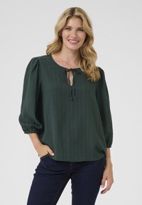 Blusa verde, strutturata, con scollo rotondo e dettaglio con nodo, caratterizzata da maniche a sbuffo a tre quarti e vestibilità rilassata. Abbinata a jeans scuri.